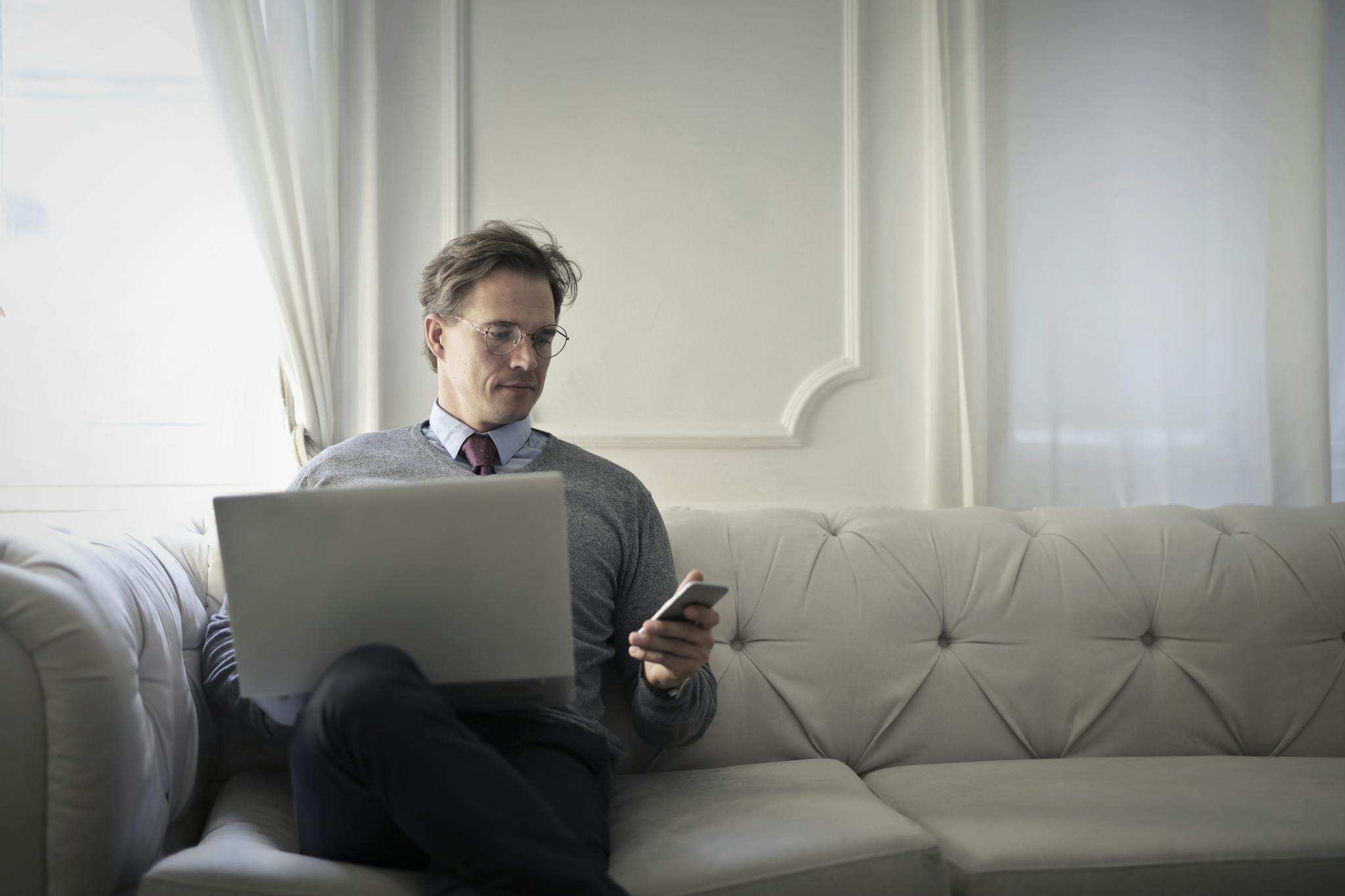 Adult man sitting on a sofa, using laptop and smartphone for remote work in a comfortable home setting.