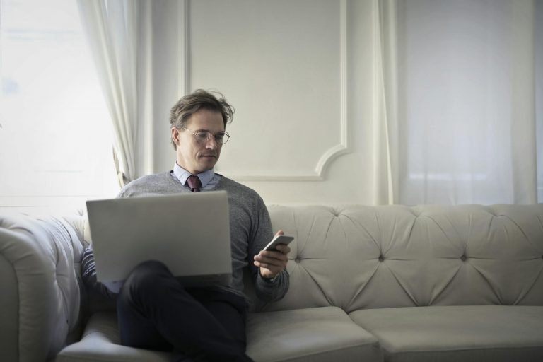 Adult man sitting on a sofa, using laptop and smartphone for remote work in a comfortable home setting.